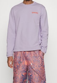 Persona che indossa un felpa a maniche lunghe color lavanda con testo arancione e pantaloni decorati con disegni floreali arancioni e blu navy.