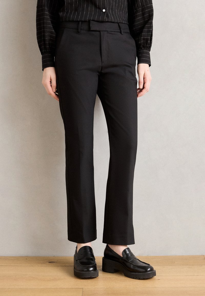 Pantalon noir sur mesure avec une coupe ajustée, devant plat et finition lisse. Associé à des mocassins noirs brillants sur un sol en bois.