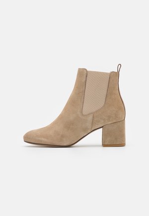 Beige ruskind ankelstøvle med blokhæle, elastisk sidepanel og trækstrop bagpå på hvid baggrund.