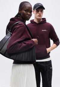 Femme en sweat à capuche bordeaux et jupe blanche portant un sac noir ; homme en sweat à capuche bordeaux et casquette noire se tient à côté d'elle sur un fond uni.