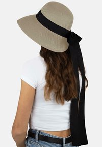 Hut mit breiter Krempe in hellbeige aus strukturiertem Stroh, mit einem schwarzen Band und einer Schleife auf der Rückseite. Getragen mit einem weißen Oberteil und blauen Jeans.