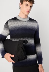 Marine- und grau gestreifter Strickpullover mit rundem Ausschnitt und strukturiertem Finish. Das Model hält eine schwarze, gepolsterte Tasche.
