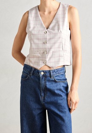 Femme portant un gilet beige sans manches boutonné avec des carreaux rose clair et un jean bleu ample taille haute.