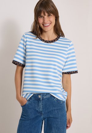 Femme souriante portant un t-shirt à manches courtes rayé bleu et blanc avec un galon imprimé léopard, et un jean bleu avec les mains dans les poches.
