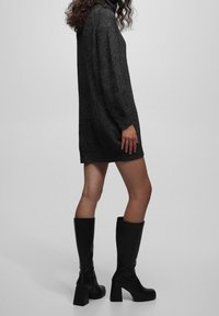 Knit, dark grey sweater dress with a high collar and loose fit. Paired with knee-high black heeled boots and a smooth texture.