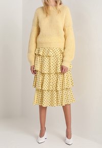 Donna che indossa un maglione giallo a maglia grossa, una gonna gialla a balze con pois neri, e scarpe con tacco bianche, in piedi davanti a uno sfondo neutro.