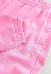 Ružové tepláky s mäkkou textúrou, elastickým pásom a prednou vreckom. Na dolnej strane sa nachádza malé biele logo "self.".