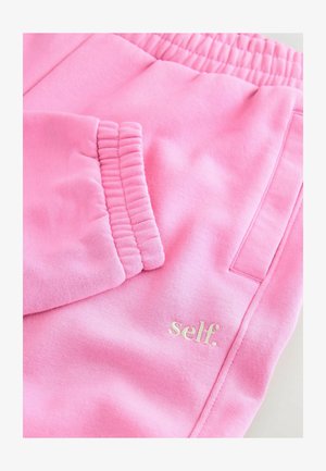 Ružičaste trenirke s mekom teksturom, elastičnim pojasom i prednjim džepom. Imaju mali bijeli logo "self." na donjoj strani.