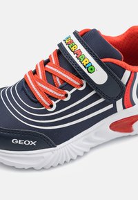 Sapatilha atlética azul-marinho com detalhes vermelhos, apresentando riscas brancas, uma faixa de velcro e marcação "Super Mario". Logotipo Geox na lateral.