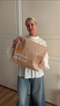 Brauner Papierbeutel mit dem Text "Love me. Wear me." in Weiß, versehen mit einer orangefarbigen Grafik. Die Person trägt eine weiße Bluse und blaue Jeans.