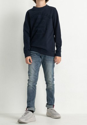 Mørkeblå sweatshirt med rund halsudskæring, med et tekstureret grafisk tryk. Kombineret med lyseblå jeans og grå sneakers.