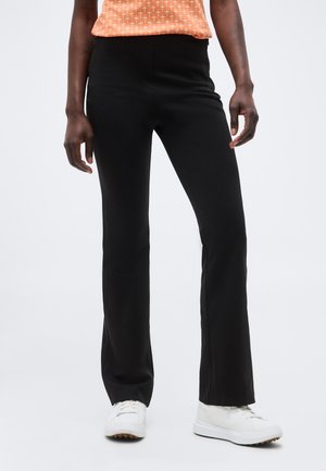 Άτομο που φοράει μαύρο παντελόνι κανονικής γραμμής, λευκά αθλητικά παπούτσια και πορτοκαλί μπλούζα με σχέδια, στέκεται μπροστά σε απλό ανοιχτό φόντο.