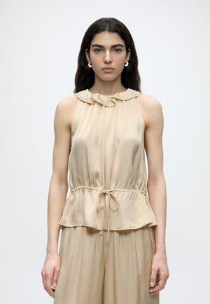 Nuori nainen, jolla on pitkät tummat hiukset, käyttää hihattoman beigen satiinitopin, jossa on röyhelökaulus ja kiristysnyöri vyötäröllä, yhdistettynä samansävyisiin housuihin.