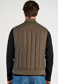 Vadderad olivgrön väst med kanalquiltning, ståkrage och ribbade mörka tröjärm, detalj med logotyp på övre delen av ryggen.