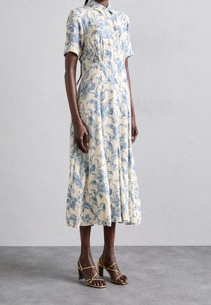 Femme portant une robe midi crème à imprimé floral bleu, manches courtes, col à revers, et sandales à talons hauts dorées à lanières sur un sol gris.