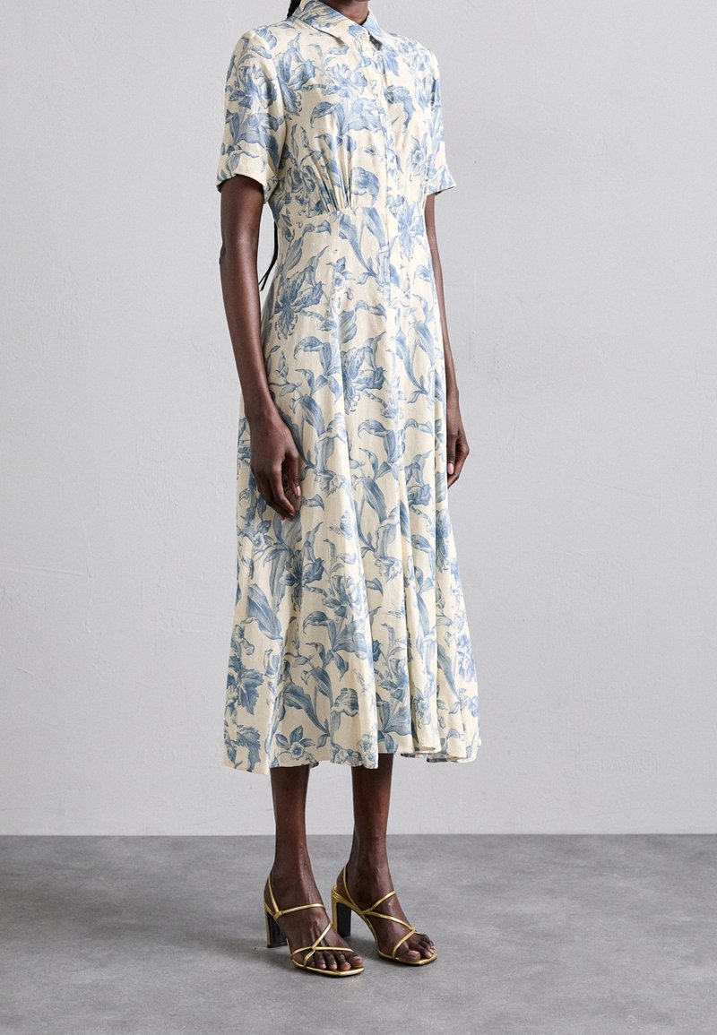 Femme portant une robe midi crème à imprimé floral bleu, manches courtes, col à revers, et sandales à talons hauts dorées à lanières sur un sol gris.