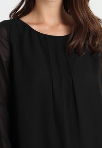 Svart blus med rundad halsringning, med genomskinliga långärmade ärmar och vertikala veck längs framtill, tillverkad av lättviktsmaterial.