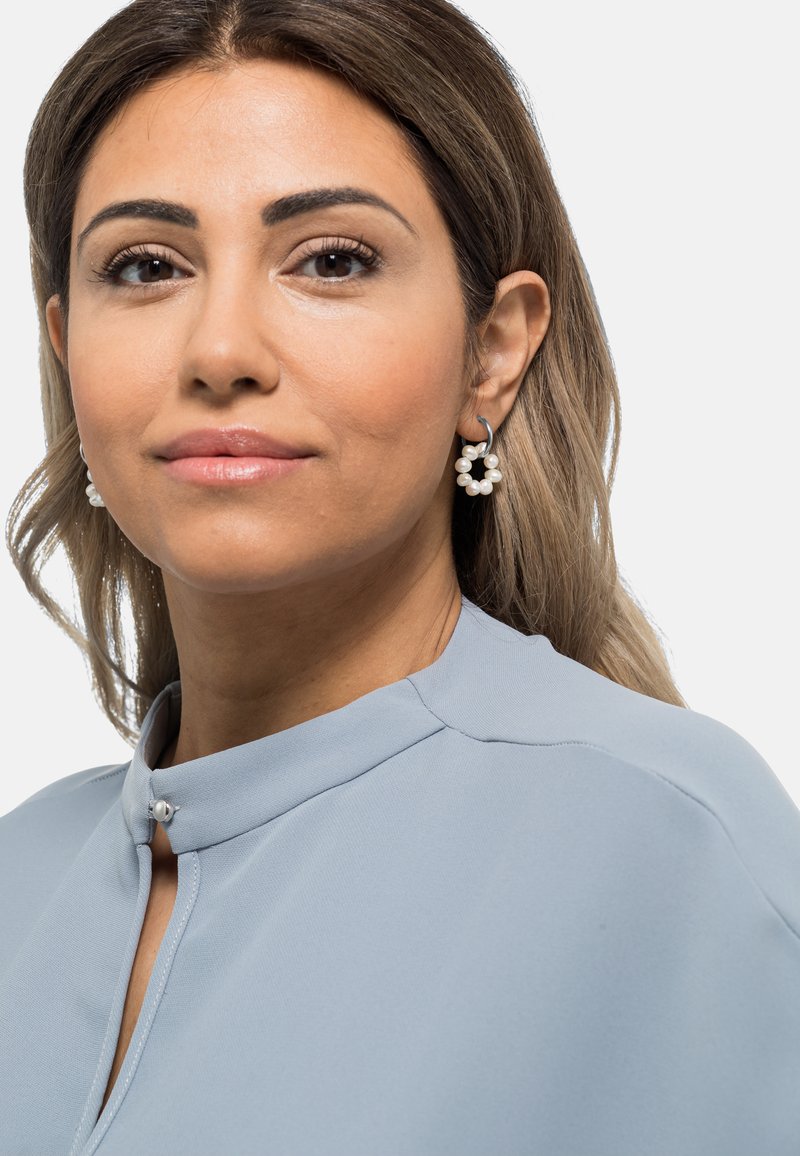 Lys blå topp med høy hals og knappedetalj. Modellen har på seg perle hoops. Glatt stoff og myk tekstur.