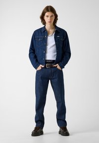 Młoda osoba stoi z rękami w kieszeniach, ubrana w ciemną kurtkę dżinsową i jeansy, biały top na ramiączkach, srebrny naszyjnik z łańcuszka oraz czarne buty.