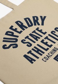 Canvas draagtas in beige met donkerblauwe opdruk. Bevat grote tekst: "SUPERDRY STATE ATHLETICS" met een coachinglabel en steraccent.