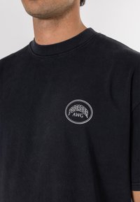 Mustmust särg, millel on ümar kaelus, ja millel on vasakul rinnal valge tikand, mis ütleb "PEGADOR AWG".