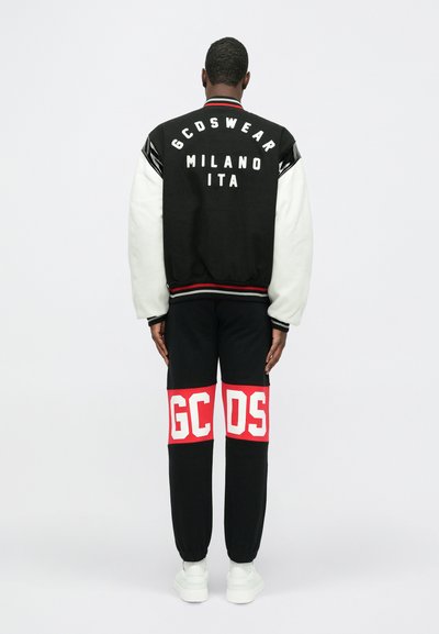 Black and white varsity jacket with logo on back, paired with black joggers featuring red and white block letters "GCDS" on thighs, and white sneakers.
