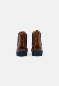 Bottes Chelsea en cuir marron avec des panneaux élastiques sur les côtés, des détails en brogue et une semelle en caoutchouc bleu marine. Présente un tirant au niveau du talon.