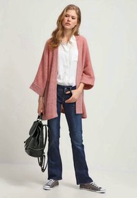 Rosa, offen gestaltete Strickjacke aus weichem Material, kombiniert mit einer weißen Bluse und blauen Jeans. Die Accessoires umfassen einen schwarzen Rucksack und graue Sneakers.