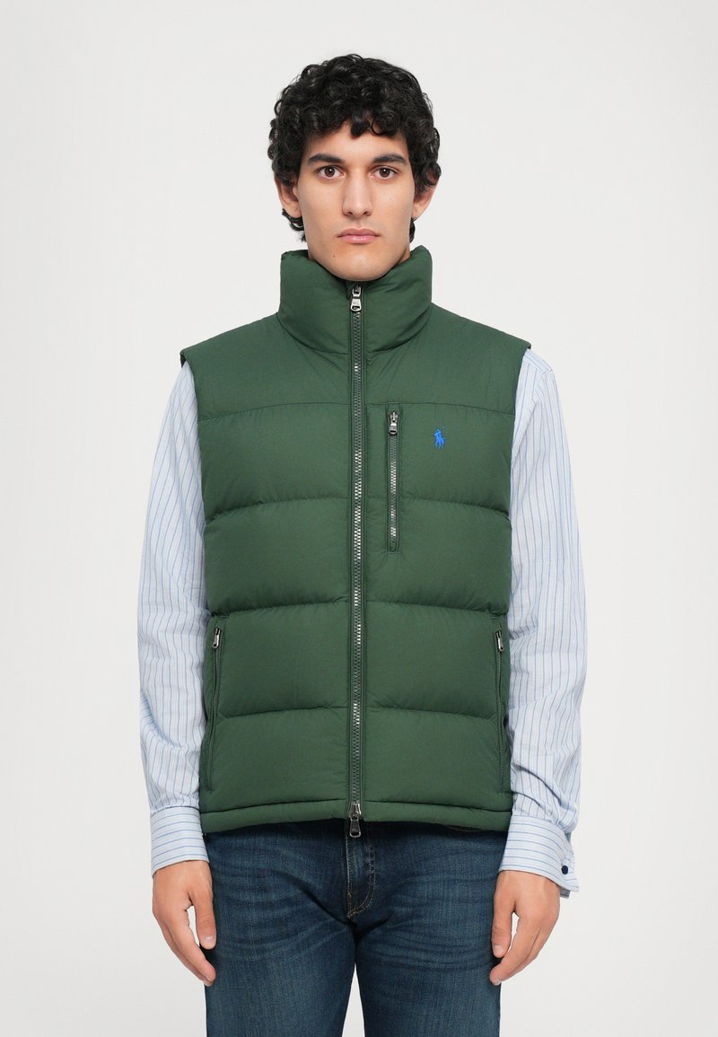 Hombre vestido con un chaleco acolchado verde oscuro sobre una camisa a rayas azul y pantalones oscuros, de pie frente a un fondo blanco liso.