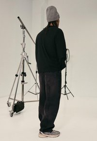 Black oversized sweatshirt with dropped shoulders, paired with black loose-fit jeans. Grey knit beanie, silver-accented sneakers. Studio backdrop.