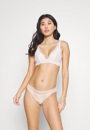 Γυναίκα με μακρύ σκούρο μαλλί, φορώντας ένα ανοιχτό ροζ δαντελένιο σουτιέν και μπεζ δαντελένια εσώρουχα, που στέκεται μπροστά σε ένα απλό λευκό φόντο.