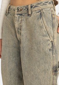 Acercamiento a unos jeans de mezclilla de talle alto y lavado claro, con bolsillos frontales, cierre de botón y detalle de tira de utilidad lateral.