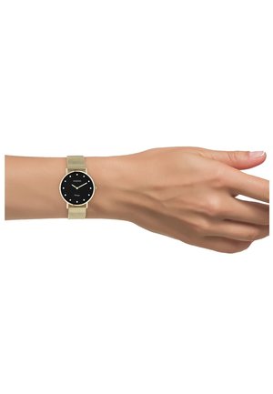 Poignet portant une montre à bracelet en maille dorée avec un cadran rond noir, des marqueurs d'heure blancs minimalistes, et des aiguilles dorées indiquant 10h10.
