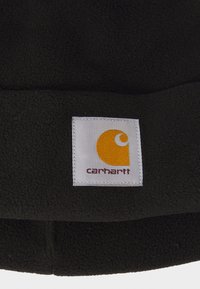 Tessuto in pile nero con un'etichetta rettangolare bianca che presenta un logo giallo e arancione. La texture appare morbida e calda.