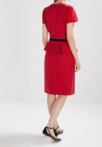 Robe peplum rouge à manches courtes et une ceinture noire, affichant une silhouette cintrée et un détail de bouton dans le dos. Portée avec des ballerines noires.