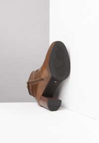 Stivale alla caviglia in pelle marrone con zip laterale, tacco impilato, suola texturizzata e finitura liscia. Posizionato su uno sfondo grigio e bianco.