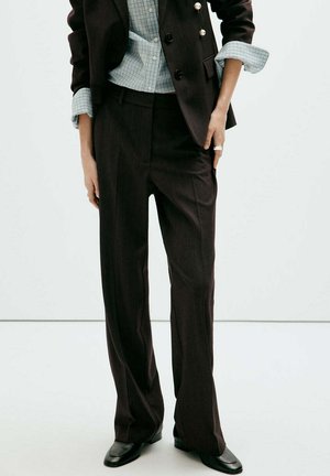 Persona que lleva pantalones negros a medida, blazer negro con botones dorados, camisa azul a cuadros con mangas remangadas y zapatos negros.