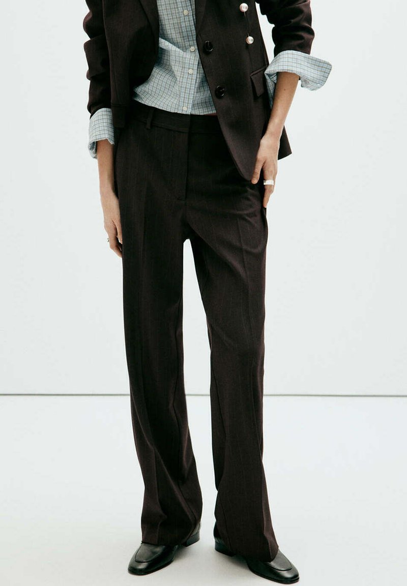 Personne portant un pantalon noir sur mesure, un blazer noir avec des boutons dorés, une chemise bleue à carreaux avec les manches retroussées et des chaussures noires.