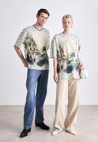 Korte ærmede grafiske t-shirts med landskabsprint i pastelfarver, båret med blå denim og cremefarvede bukser, suppleret af sort- og hvidt fodtøj.