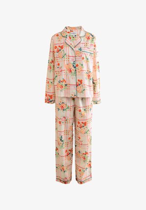 Kvinde stående i blomsterprintede pyjamas i et blødt oplyst rum med ferskenfarvede vægge og blondegardiner over to vinduer.