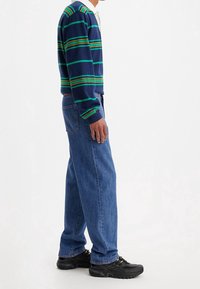 Námořnický pruhovaný svetr se zelenými a žlutými pruhy, v kombinaci se světle modrými džíny rovného střihu a černými teniskami s nízkým profilem.