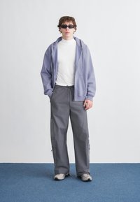 Casaco com capuz roxo claro com fecho de correr, gola alta branca, calças cargo cinzentas com fechos de correr e ténis cinzentos. Os acessórios incluem óculos de sol retangulares pretos.