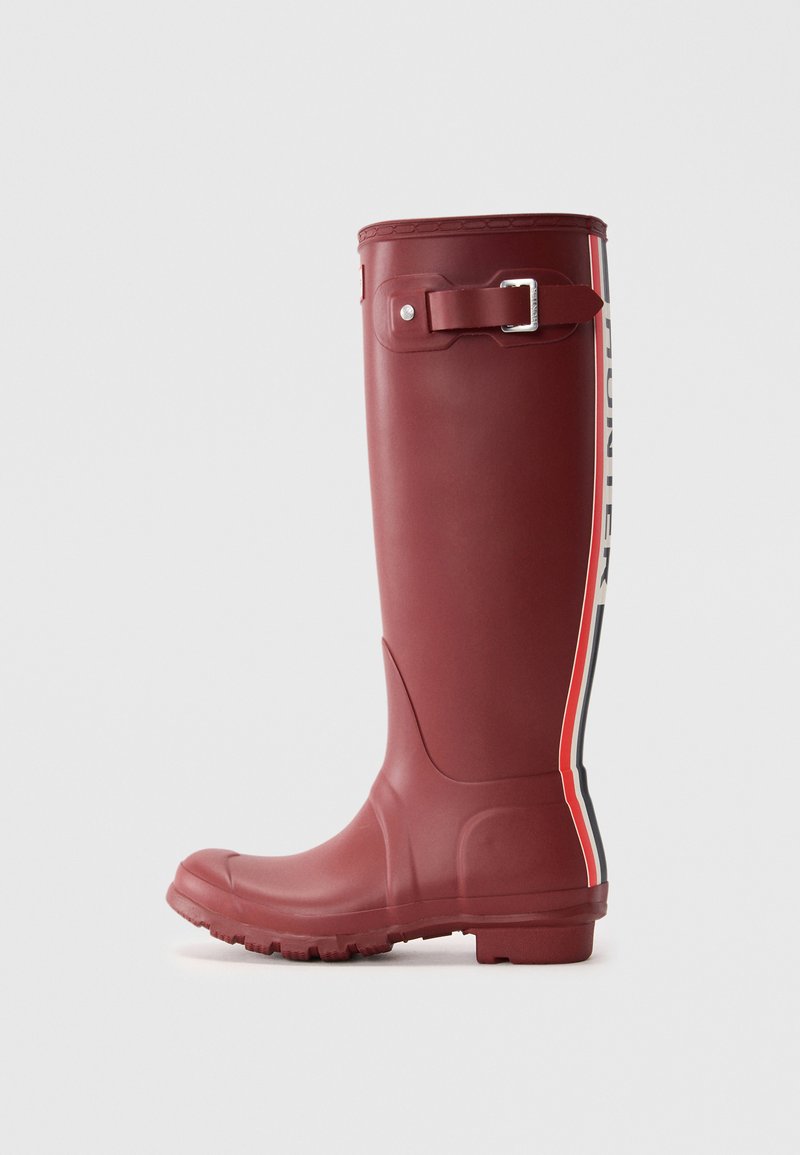 Botte en caoutchouc bordeaux hauteur genou avec une boucle sur le côté, surface texturée, et une bande contrastante blanche et rouge portant le nom de la marque.