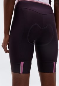 Κοντινό πλάνο ενός ατόμου που φορά μαύρο ποδηλατικό σορτς με ροζ λεπτομέρειες και επενδυμένη ραφή στην περιοχή της καρέκλας, στέκεται μπροστά από απλό φόντο.