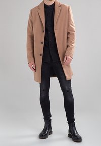 Homme portant un manteau beige sur une chemise noire, un jean skinny noir déchiré et des bottes en cuir noires, se tenant devant un fond uni.