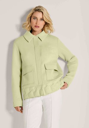 Blonde Frau trägt eine hellgrüne Jacke mit großen Taschen und einem Rüschensaum, kombiniert mit weißen Hosen, steht vor einem schlichten Hintergrund.