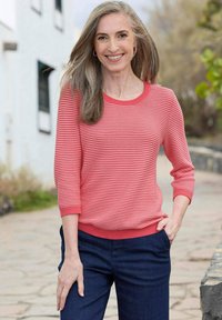Koralfarben gestreifter Pullover mit drei Viertel-Ärmeln und geribtem Kragen, kombiniert mit dunklen, geraden Hosen. Glatte Stofftextur sichtbar.