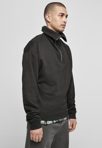 Sweatshirt negru, cu guler înalt și jumătate de fermoar, fabricat dintr-un material moale, având manșete ribbate și o croială confortabilă.