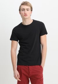 Camiseta de algodón negra con cuello redondo y mangas cortas, combinada con pantalones rojos, presentada sobre un fondo liso.