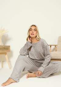 Kvinna sitter på vit golv iklädd grå texturerad loungedräkt, med ett sidobord i ljus trä och en beige stol i ett minimalistiskt rum.